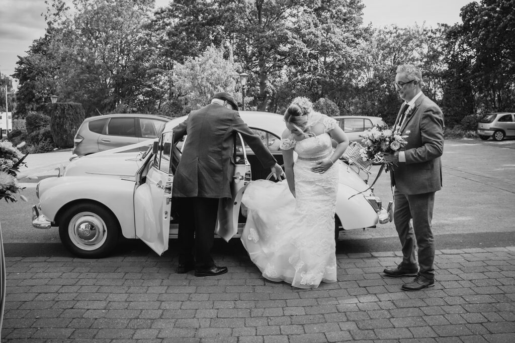 Bride exiting vintage car with groom and groomsman at wedding