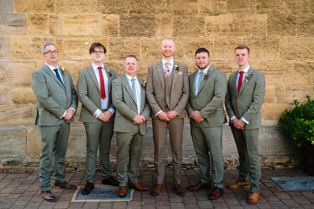 Six men in formal suits standing against stone wall backdrop