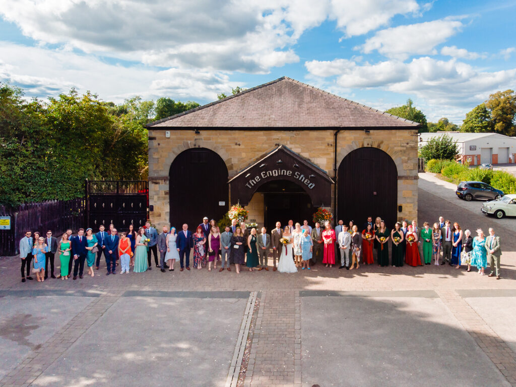 Large wedding party posed outside historic stone engine shed building