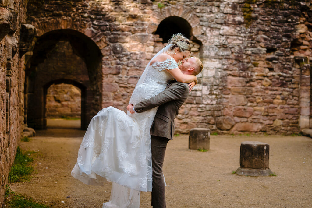 Groom lifting bride in white dress at historic stone ruins