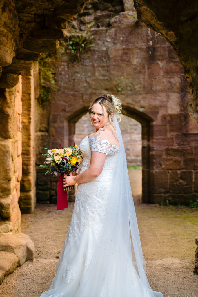 Bride in white lace dress holding colourful bouquet at historic stone ruins