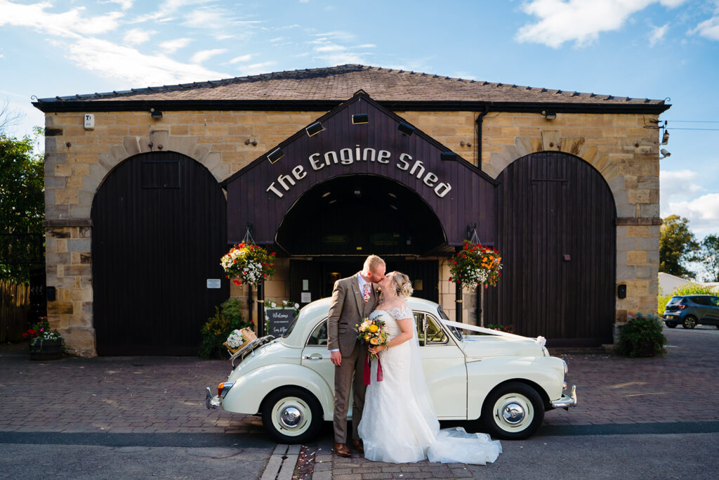 Bride and groom kissing beside vintage white car at Engine Shed
