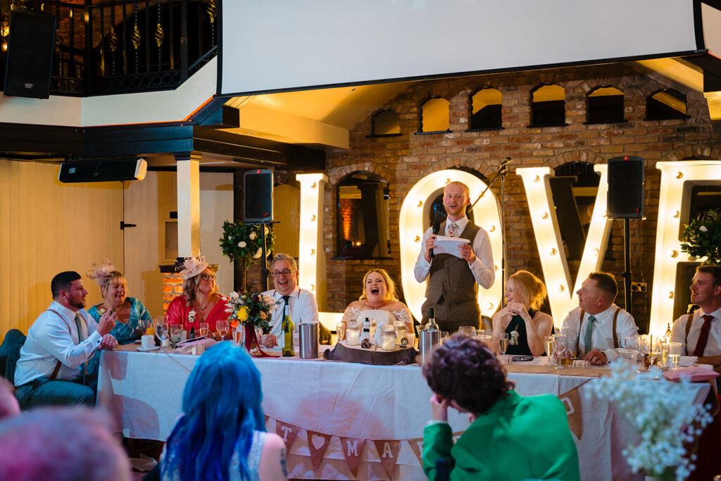 Best man giving speech at wedding reception with illuminated LOVE sign