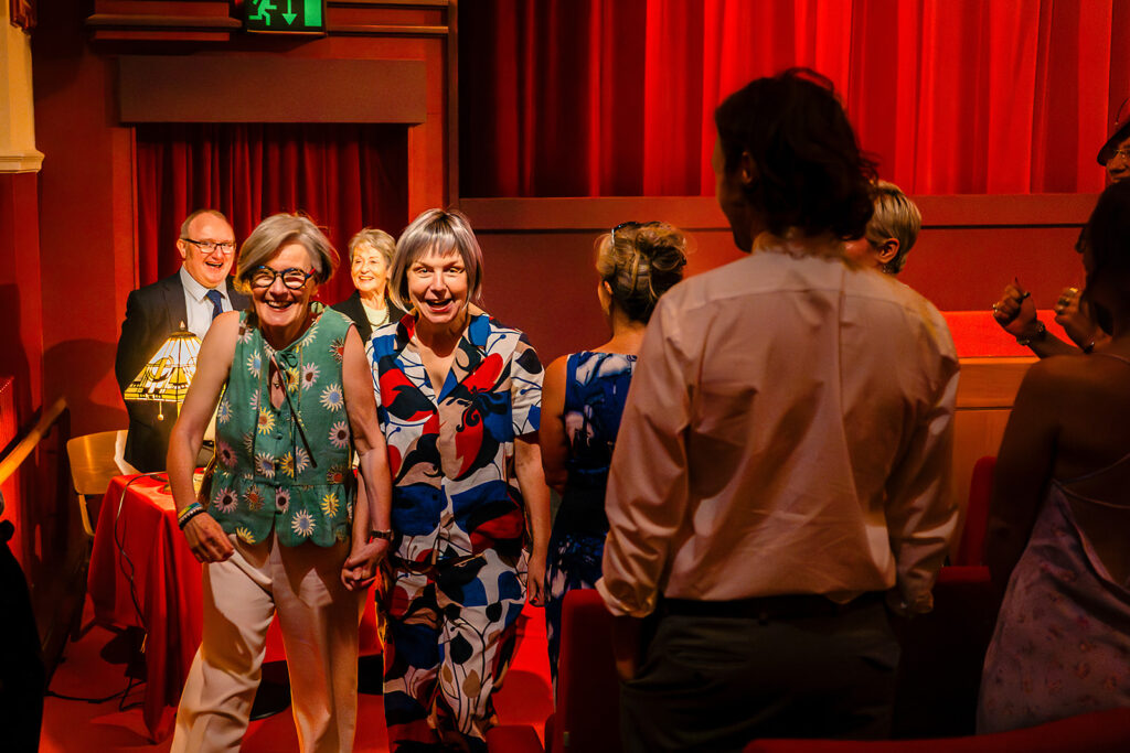 Beverly and Jane walking into the red auditorium of the Hyde Park Picture House together, smiling at their guests