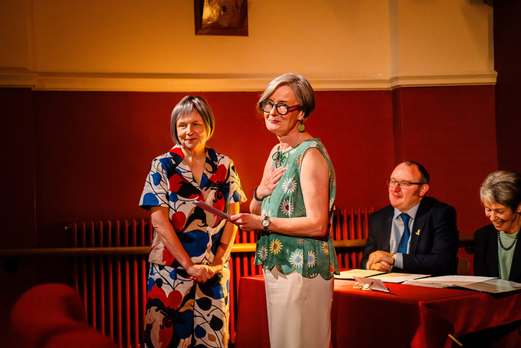 Beverly and Jane standing hand on heart at the front of the auditorium, with the registrars at their table