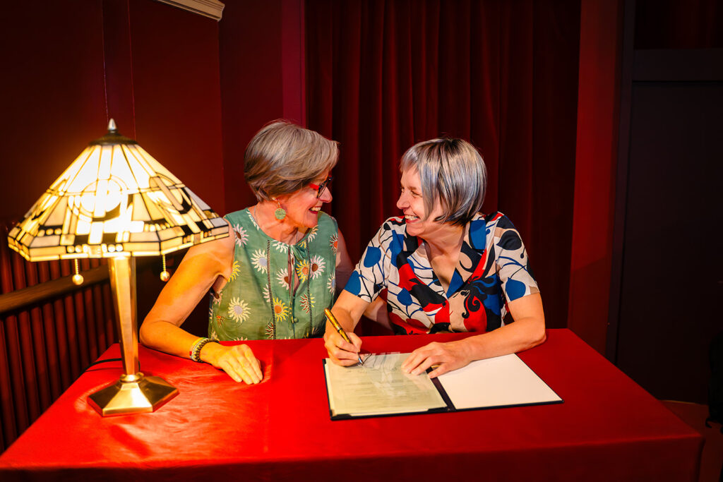 Beverly and Jane signing the register at a red table under a Tiffany-style lamp