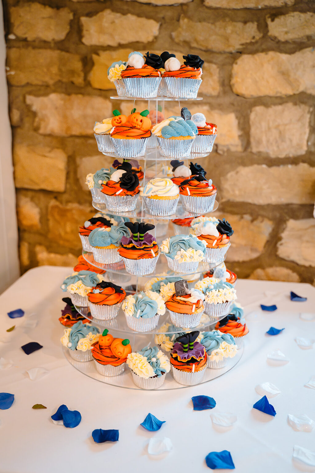Tower of Halloween wedding cupcakes in orange, blue and black on a table scattered with petals