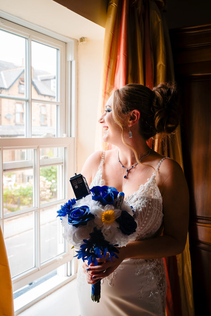 Bride in white dress holding blue bouquet by window