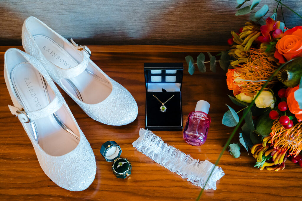 White bridal shoes, wedding rings, a green pendant necklace, perfume and a garter laid out next to an orange bouquet
