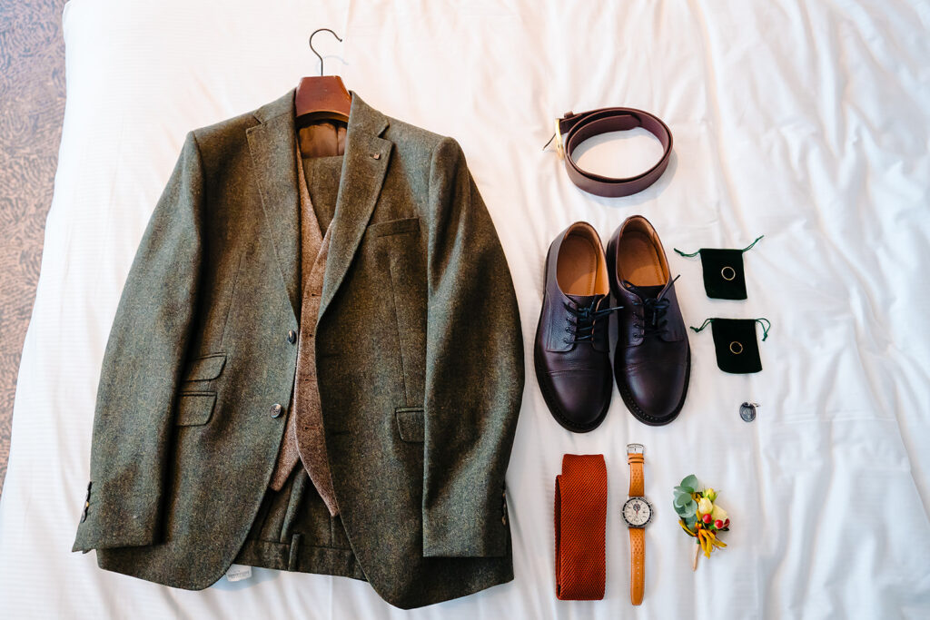 Groom's green tweed three-piece suit laid out with brown shoes, a burnt-orange knitted tie, a watch and a buttonhole