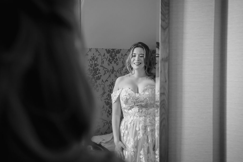 Black and white photo of Ellissa smiling in her floral off-the-shoulder wedding dress in a hotel doorway