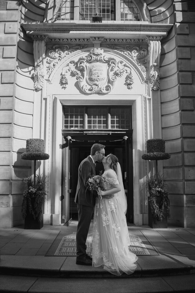 Black and white photo of Ellissa and Anthony kissing in front of an ornate carved stone doorway in York