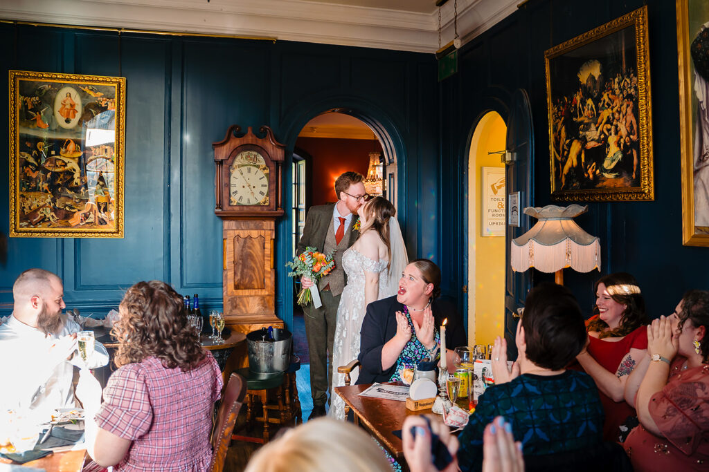 Guests cheering as Ellissa and Anthony kiss in the navy-panelled Blue Room at the House of the Trembling Madness, York