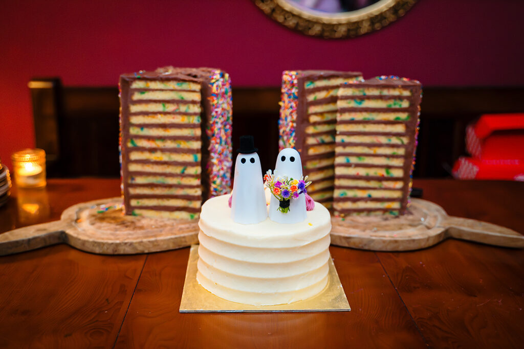 Small white wedding cake with two ghost cake toppers, one in a top hat and one holding a bouquet, in front of tall sprinkle layer cakes