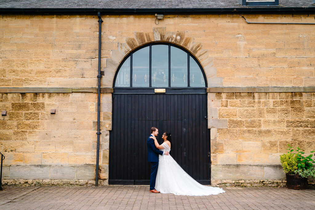 Fran and Andy holding each other in front of a tall black arched door on the stone side of the Engine Shed