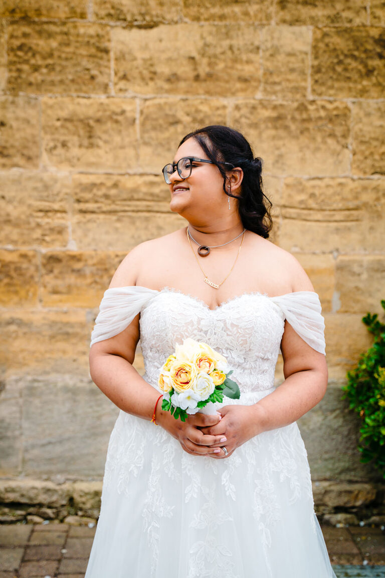 Fran smiling and looking away, holding her yellow rose bouquet against the Engine Shed's stone wall