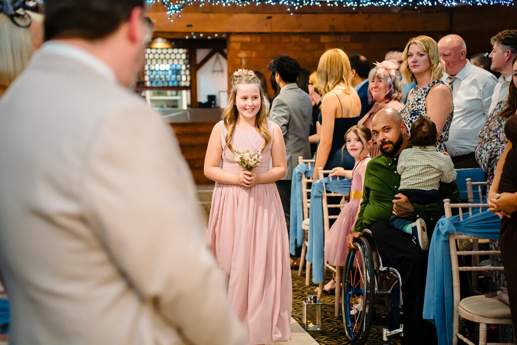A flower girl in pink walking down the aisle of the Horseshoe Barn as guests turn to watch