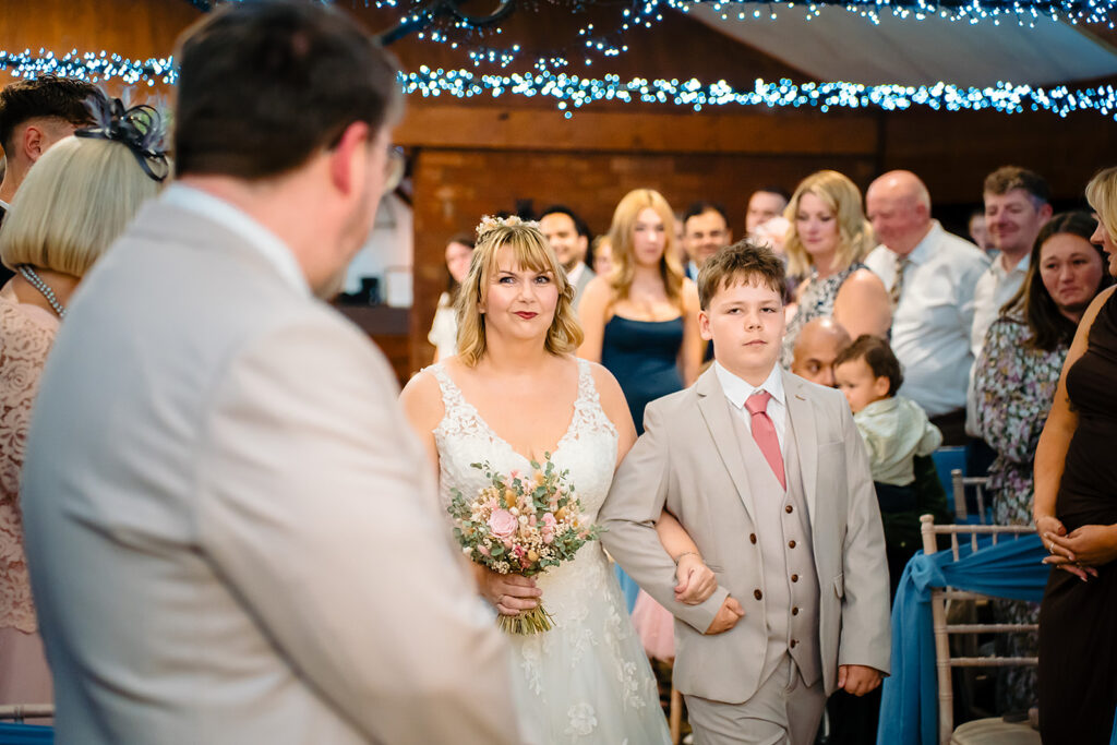 Kay walking down the aisle with their son as Steven waits at the front of the Horseshoe Barn