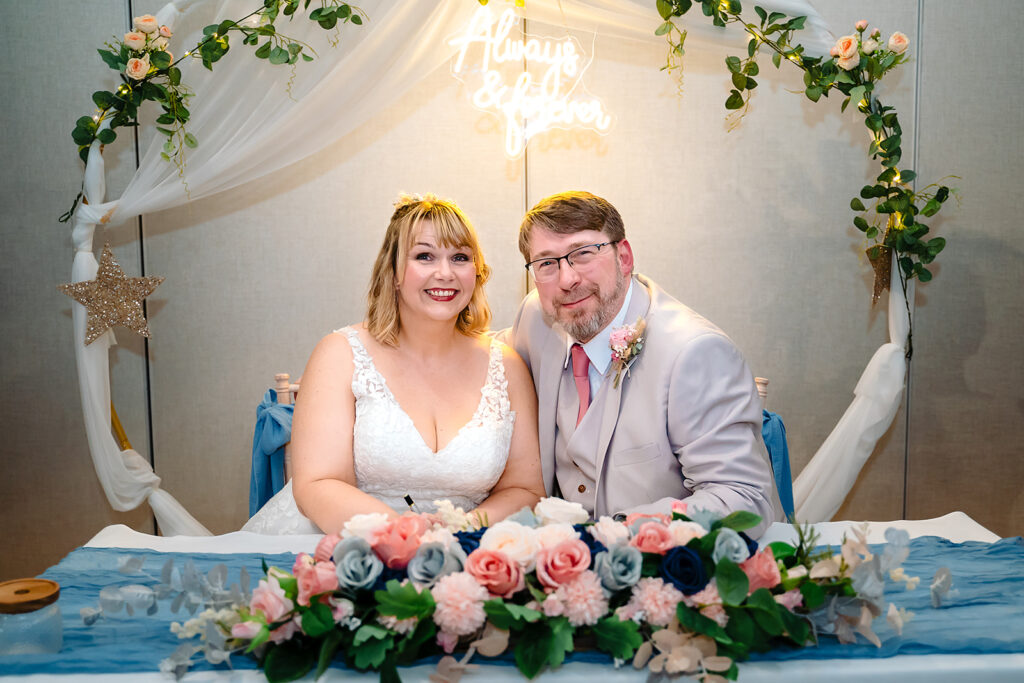 Kay and Steven smiling at the signing table beneath an Always and Forever neon sign