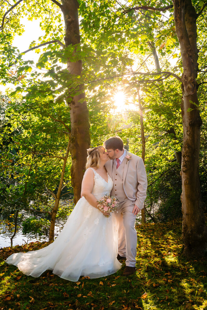 Kay and Steven embracing under the trees with the sun shining through the branches behind them