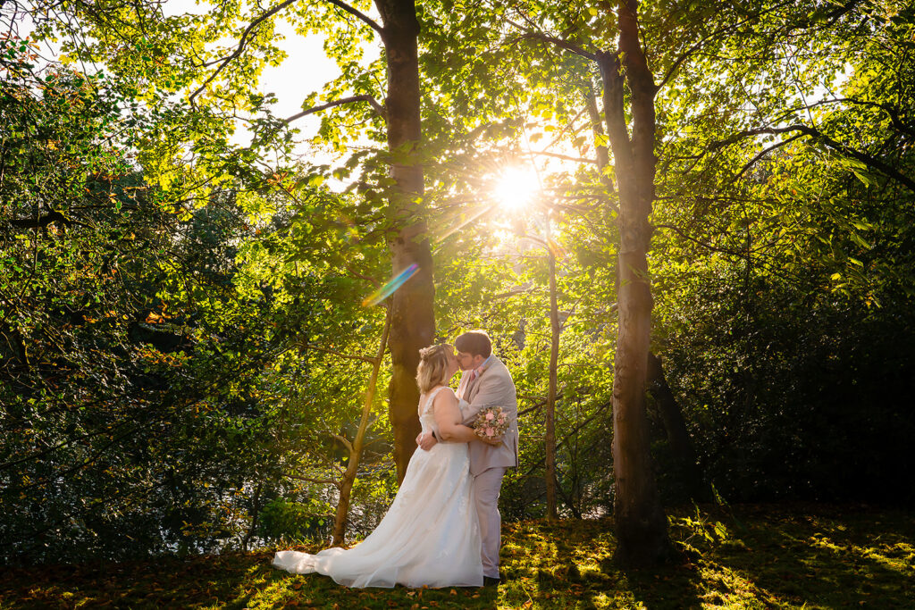 Kay and Steven kissing among the trees as the low sun bursts through the leaves at the Stables Country Club, Bury