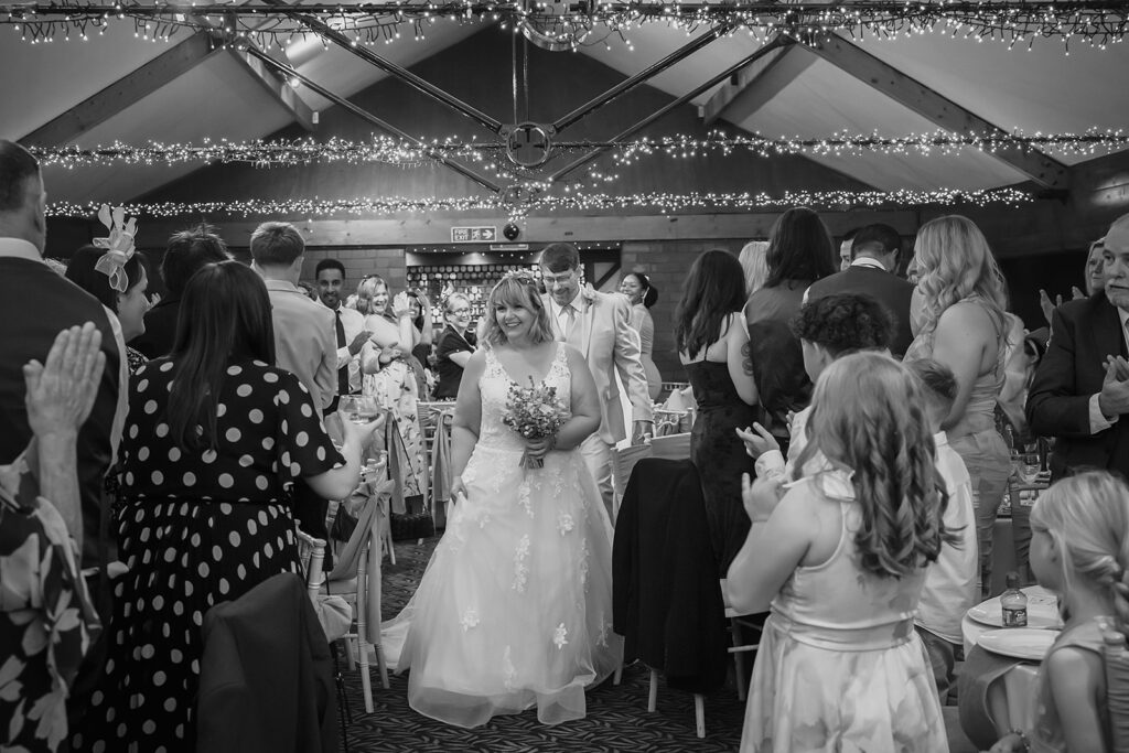 Black and white photo of Kay walking into the reception as guests stand and clap