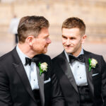 Two grooms in black tuxedos smiling at each other.