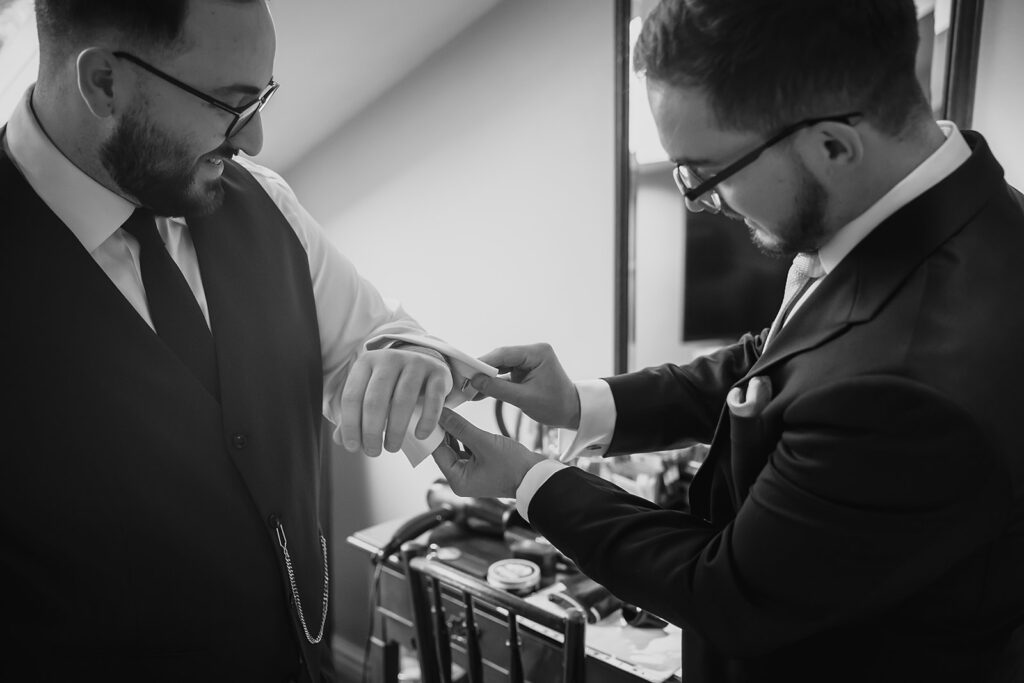 Black and white photo of a groomsman helping Nicholas with his cufflinks on the wedding morning