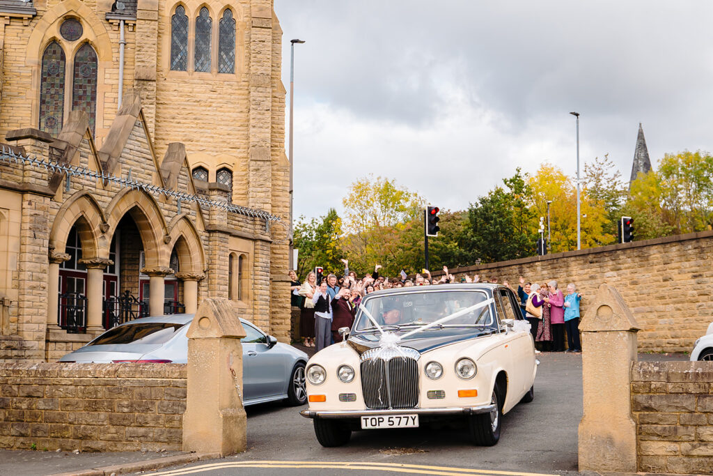 Guests waving as the vintage wedding car pulls away from Dewsbury Baptist Church