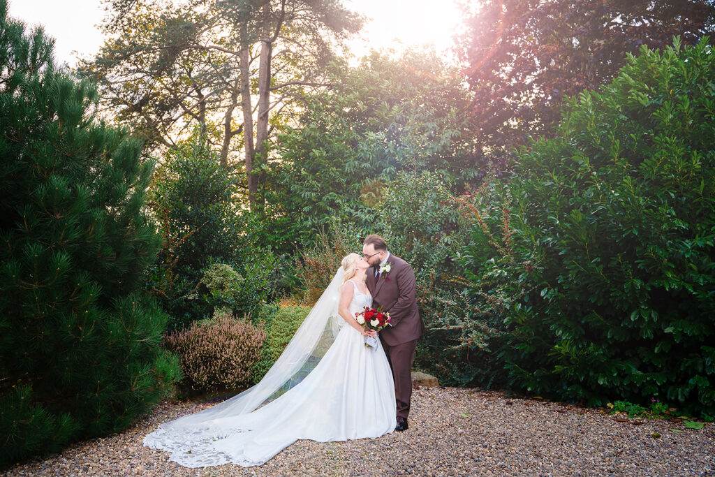 Rebecca and Nicholas kissing in the sunlit gardens at Holdsworth House, Halifax, with her long veil spread on the gravel