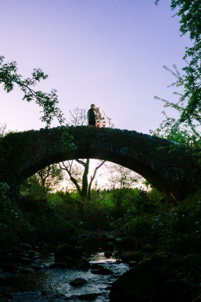 Couple atop stone bridge at sunset, lanterns glowing near The Out Barn.